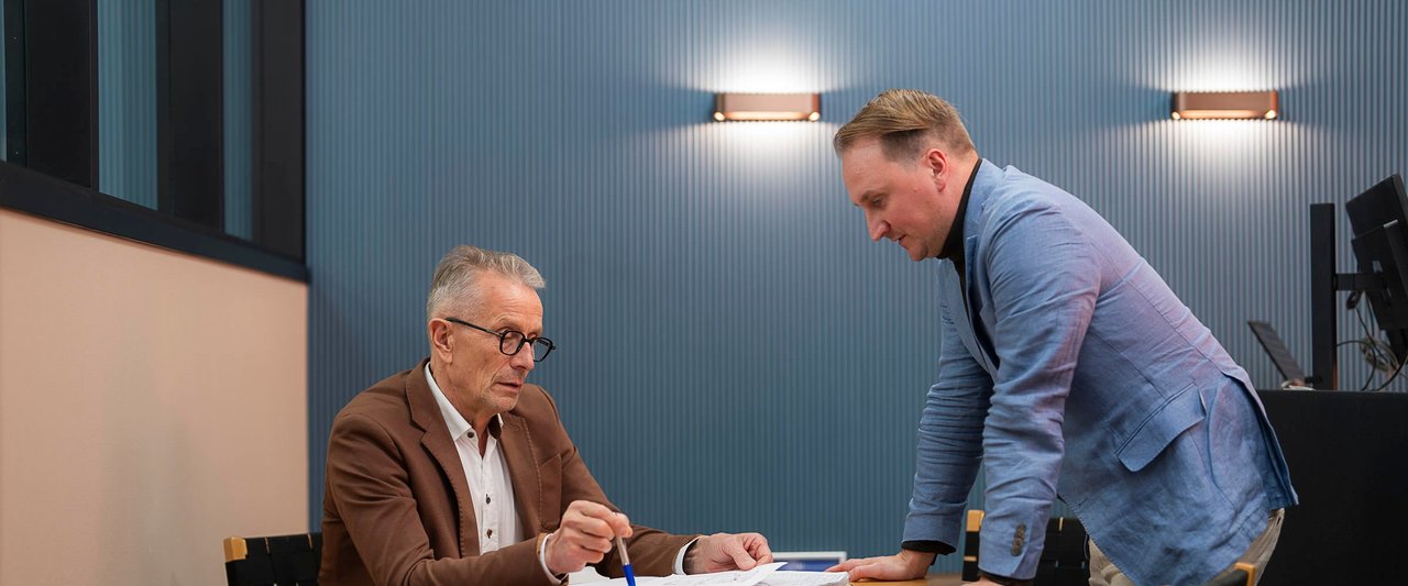 Nainen istuu villaneuleessa nojatuolissa suuren ikkunan ääressä ja selaa puhelinta talvisena päivänä, vieressään kynttilälyhty ja kahvimuki.